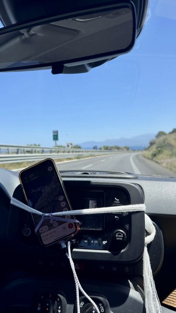 Ein Auto von innen. An der Armatur hängt ein Handy das mit einer weißen Schnur fest gespachtelt wurde durch die Windschutzscheibe sieht man das Meer und Berge