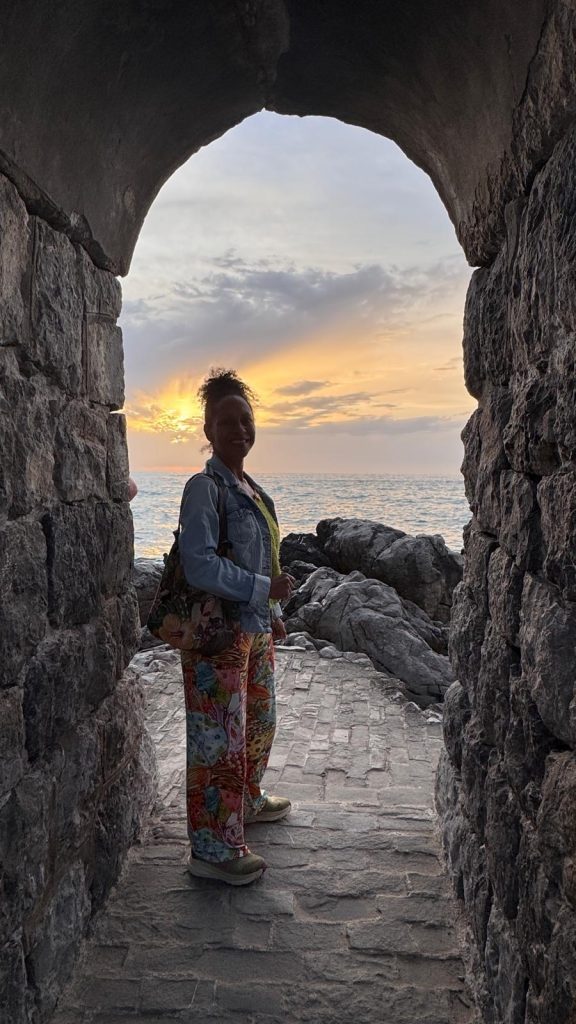 Eine Frau steht in Cefalu am Meer mit links und rechts Mauern. Im Hintergrund Sonnenuntergang