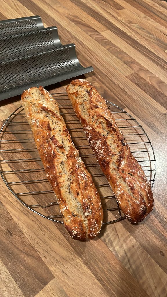 Zwei frisch gebackene Baguette, liegen auf einem runden Gitter zum abkühlen. Dahinter liegt die Baguetteform