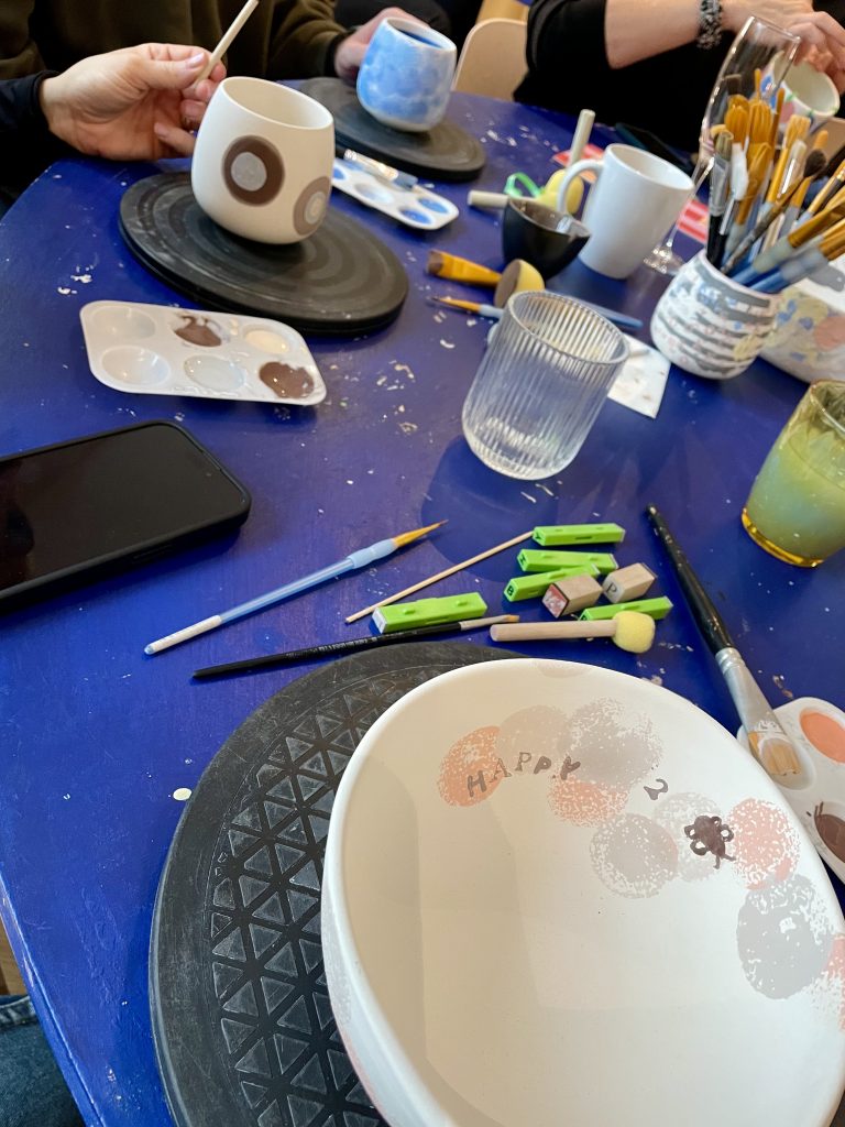 Keramikmal-Workshop mit mehreren bemalten Tassen und Schalen auf einem Tisch, umgeben von Pinseln, Farben und Werkzeugen in gemütlicher Runde.