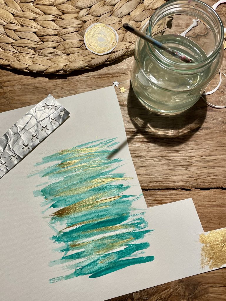 Ein Blatt Papier auf das grüne und goldfarbene Striche mit Aquarellfarben gemalt wurden. Ein Wasserglas mit einem Pinsel darin steht oberhalb des Papiers. Es werden Weihnachtskarten mit Upcycling gebastelt