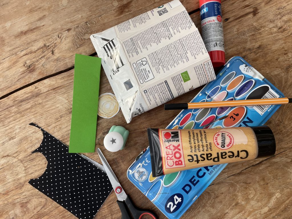 Ausgebreitetes Bastelmaterial auf einem Holztisch: ein aufgeschnittener Tetrapack, ein grüner Papierstreifen, ein Stück schwarzer Stoff mit weißen Punkten, eine Schere, ein Stern-Ausstanzer, eine Rolle Klebeband, ein Klebestift, ein Aquarellkasten, ein schwarzer Stift und eine Tube goldene Acryl-Effektpaste.