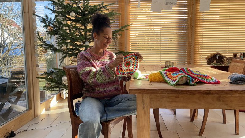 Ein Frau sitzt an einem Holztisch und häkelt bunte Gerannt Square
