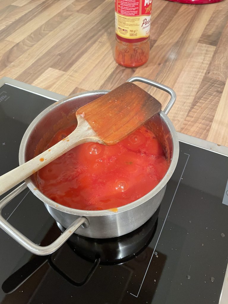 Ein Topf mit Tomatensoße steht auf dem Herd und auf dem Rand des Topfs liegt eine Kochlöffel