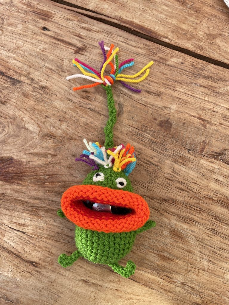 Kleine gehäkelte grüne Figur mit großen orangefarbenen Lippen und bunten Wollhaaren auf einem Holztisch Man nennt es Schlüsselfresserchen.