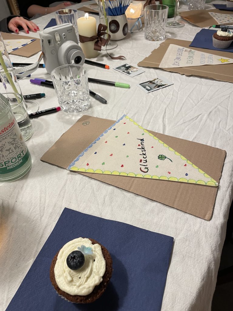 Ein Tisch auf dem Gläser stehen, eine Kamera, Farbstifte und bemalte Dreiecke aus Stoff.