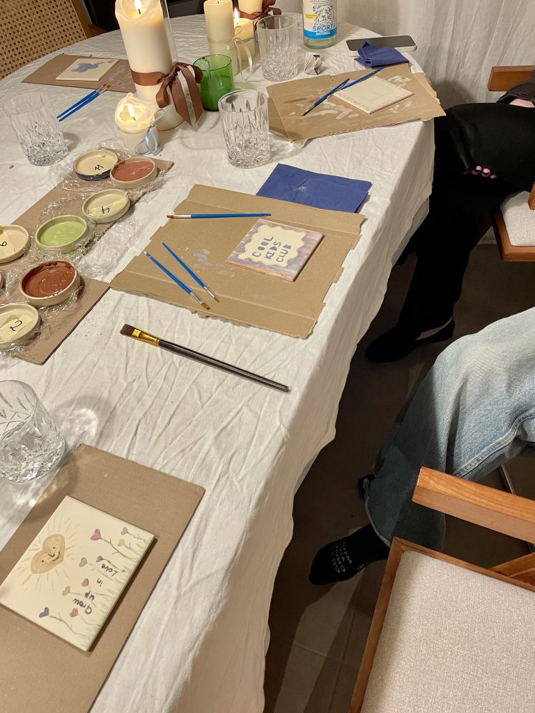 Bemalte Kacheln liegen auf einem Tisch.In der Mitte des Tischs sind Töpfchen mit Farben darin. Kerzen stehen auf dem Tisch