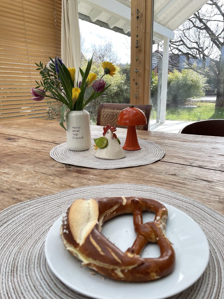 Eine Butterbrezel auf einem Teller der auf einem Holztisch steht. davor eine Vase mit Blumen, eine getöpfertes Huhn und eine oragngefarbene Tischlampe