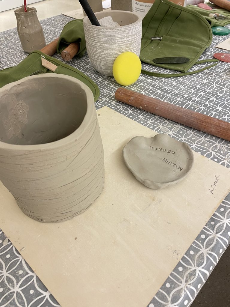 frisch getöpferte Gefäße auf einem Holzbrett. Daneben liegen Utensilien die zum töpfern gebraucht werden.