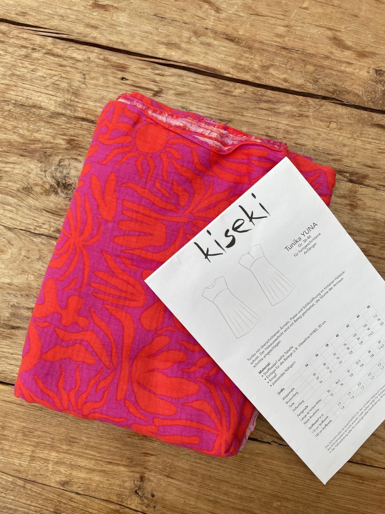 Ein leuchtend Oragne/pink gemusterter Stoff liegt zusammengefaltet auf einem Holztisch. Auf dem Stoff liegt noch ein Schnittmuster, auf dem "Kiseki" darauf steht