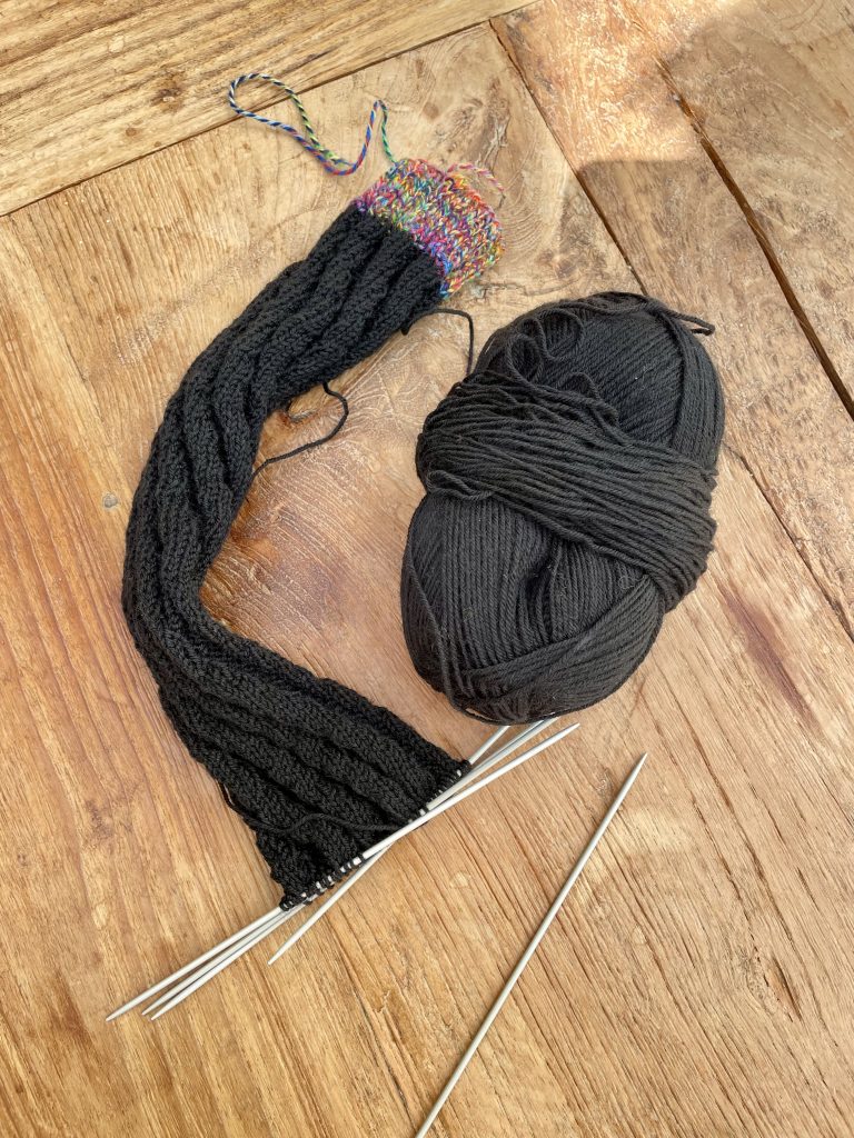 ein halb fertig gestricker Socke mit den Stricknadeln in den Maschen und dem Wollknäuel daneben.