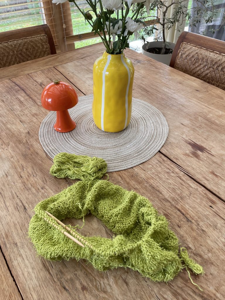 Auf einem Holztisch liegt Strickzeug. Daneben ist ein rundes Tischset auf dem eine kleine orangefarbene Lampe und eine gelb, weiß gestreifte Vase mit weißen Blumen darin steht