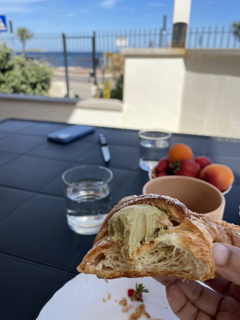 Ein italienisches Cornetto mit Pistaziencreme gefüllt angebissen. Im Hintergrund sieht man das Meer
