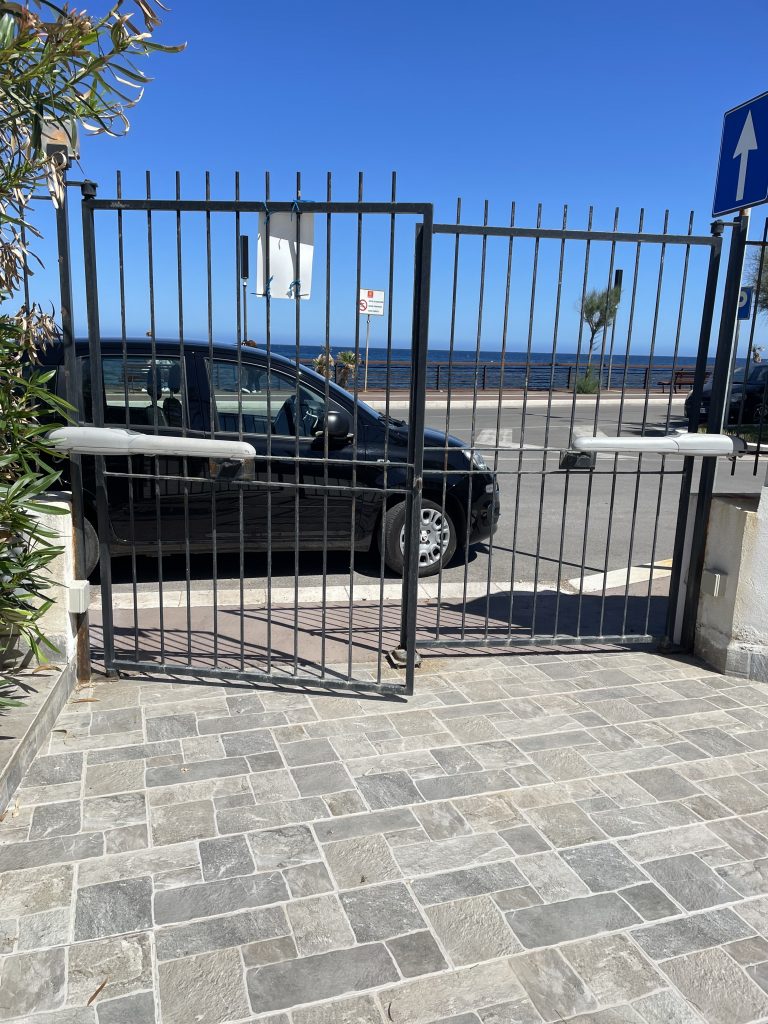 Ein halb geöffnetes Hoftor mit Meer und Strandpromenade im Hintergrung