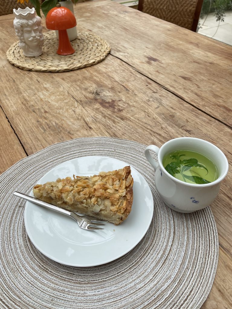 Ein Stück Kuchen auf einem Teller der auf einem Holztisch steht. Daneben eine Tasse Kräutertee