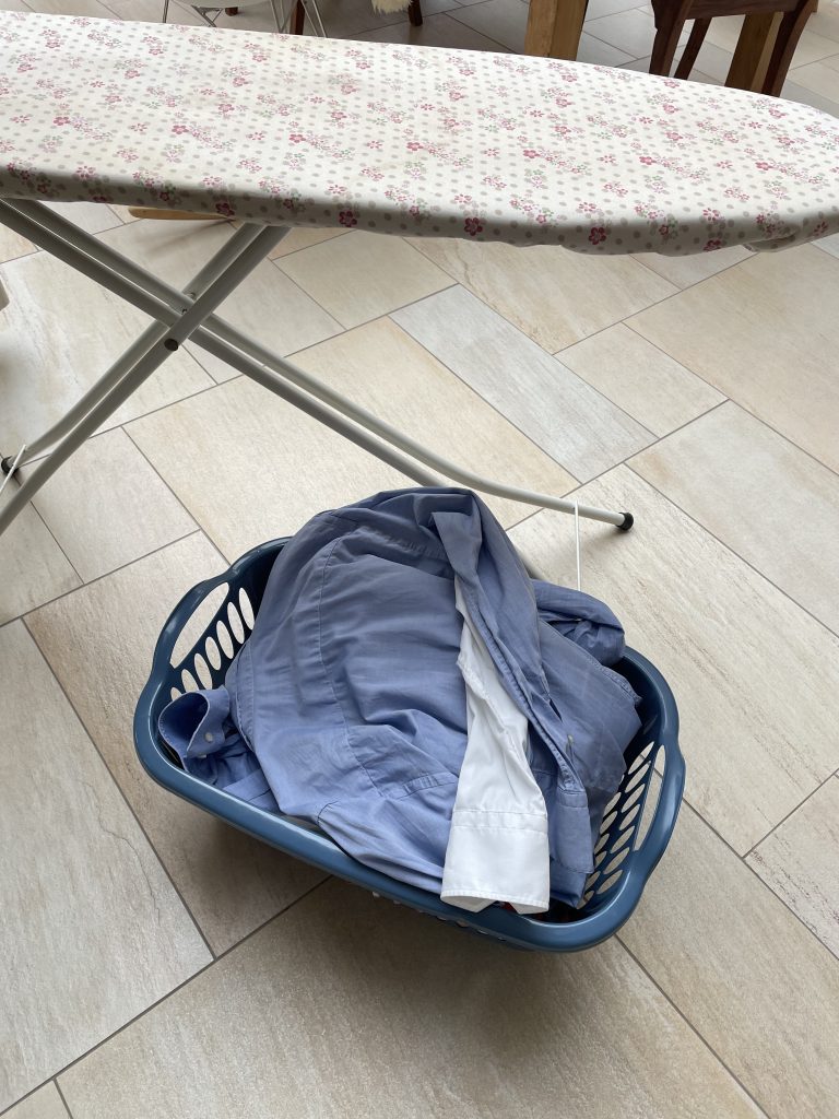 Ein Bügelbrett steht auf einem Fliesenboden und eine gefüllter Wäschekorb daneben.