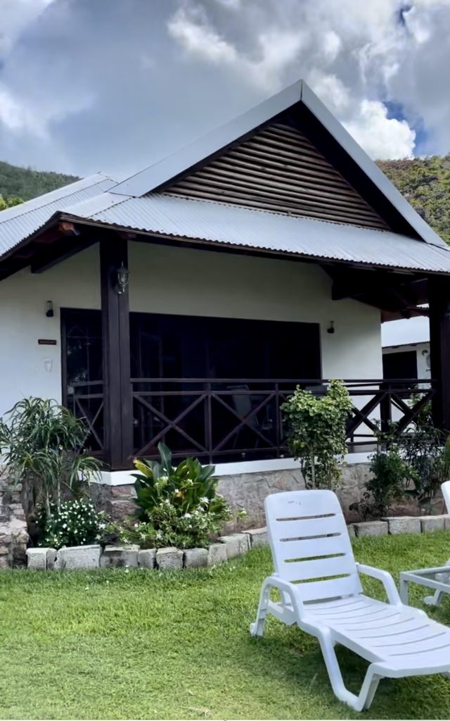 Ein Haus auf Praslin, Seychellen. Im Giebel sin vertikale schmale. Seit führen 2 schwarze Balken hinunter auf die Terrasse, deren Geländer besteht aus schwarzen gekreuzten dünnen Balken. Vor der Terrasse ist ein Pfalneznebeet, davor eine Wiese mit einem weißen Liegestuhl. Der Himmel ist bewölkt.