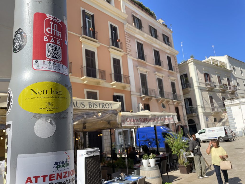 Ein Laternepfahl in Bari auf dem ein Aufkleber mit dem Text "Nett hier, aber warst du schon mal in Baden Württemberg" klebt. Im Hintergrund Gebäude und blauer Himmel