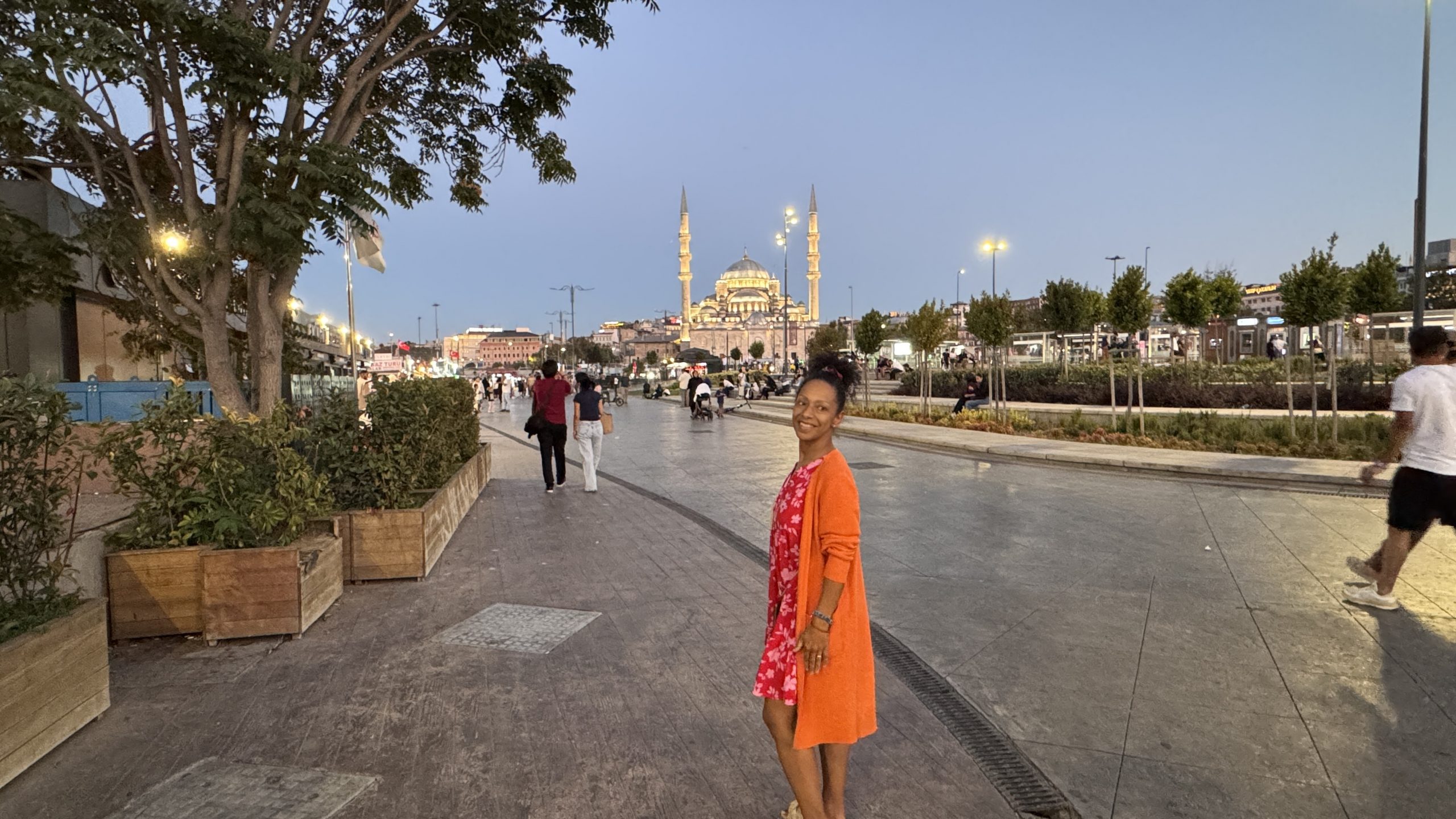 Frau in rotem Kleid mit orangefarbener Jacke steht lächelnd am Abend in Istanbul, im Hintergrund die beleuchtete Yeni Cami Moschee und eine belebte Promenade