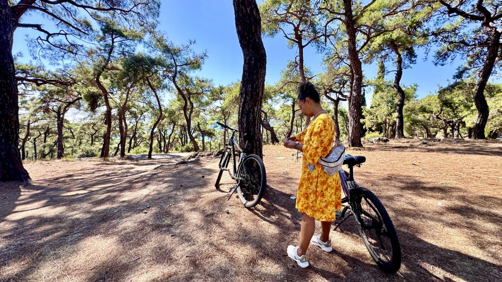 Eine Frau i einem gelben Kleid steht in ein lichten Wald neben einem Fahrrad. Vor ihr ma Baum lehnt steht noch ein anderes Fahrrad.