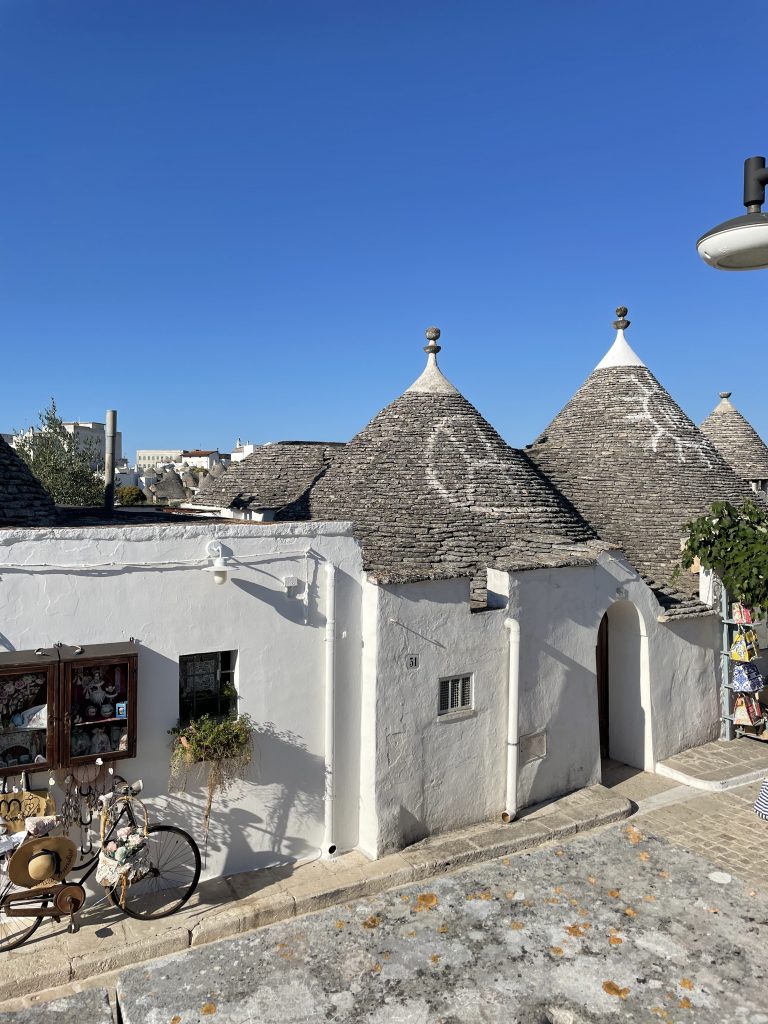 Traditionelle Trulli-Häuser mit weiß getünchten Wänden und kegelförmigen Steindächern in Alberobello, Apulien, vor strahlend blauem Himmel, im Vordergrund ein dekoriertes Fahrrad mit Blumen vor einem Souvenirladen