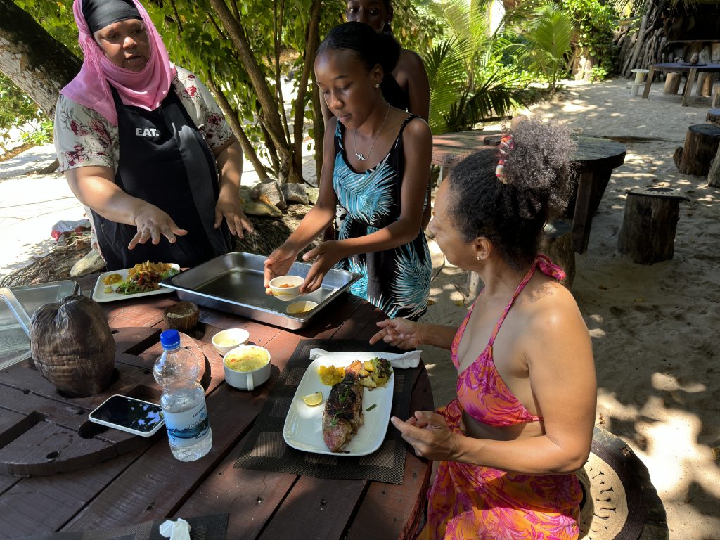 Auf den Seychellen, Eine Frau, gekleidet mit einem Orange-pinkfarbenen Bikinioberteil und einem gleichfarbigen Tuch um die Hüften,sitzt am Tisch. Sie wird von einer älteren Frau und einem jungen Mädchen bedient.Vor der Frau steht ein Teller mit gegrillten Fisch. das ganze ist an einem Strand