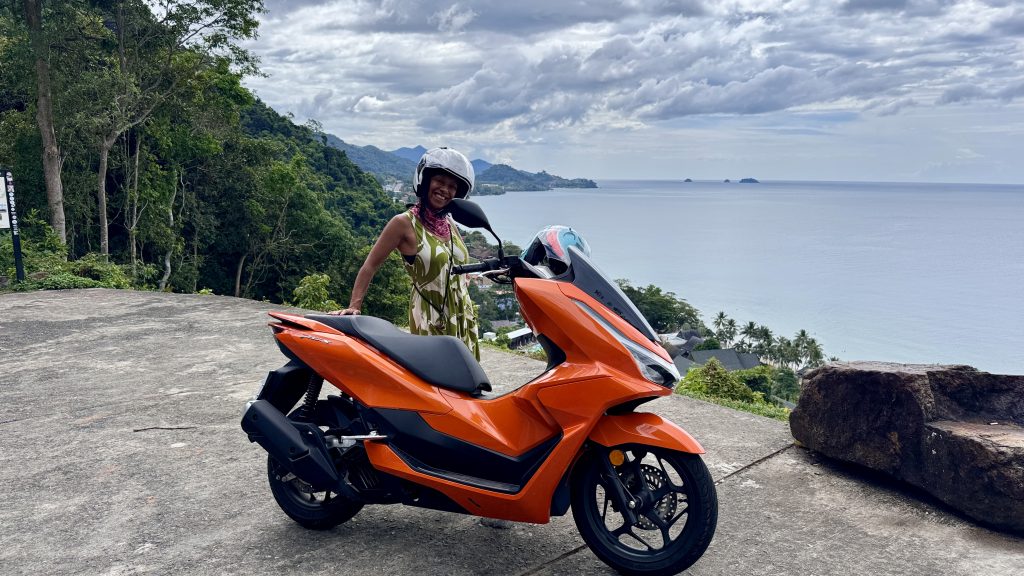 Eine Frau steht neben einem orangefarbene Motorroller auf einer Aussichtsplattform auf Koh Chang. Im Hintergrund sieht man das Meer und kleine Inseln