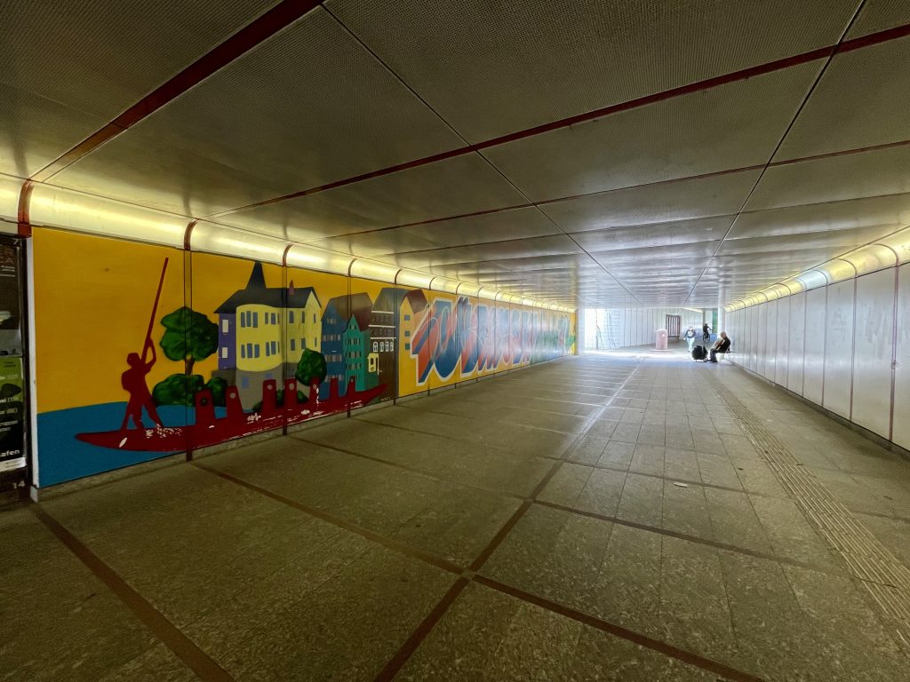 Unterführung am Hauptbahnhof Tübingen mit bunter Wandmalerei: Graffiti zeigt Stocherkahn, Altstadthäuser und Schriftzug „Tübingen“, modernes Stadtmotiv mit Personen im Hintergrund.