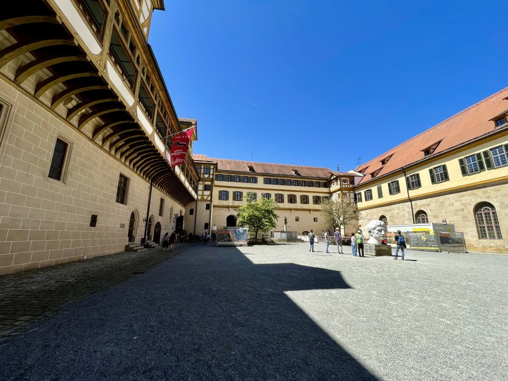 Innenhof von Schloss Hohentübingen an einem sonnigen Tag mit klarblauem Himmel. Besucher spazieren über den weitläufigen Platz, im Vordergrund ist eine große Skulptur zu sehen. Das imposante Renaissance-Gebäude beherbergt heute Teile des Museums der Universität Tübingen (MUT) mit archäologischen und wissenschaftlichen Sammlungen. Sehenswürdigkeit in Tübingen, Baden-Württemberg.