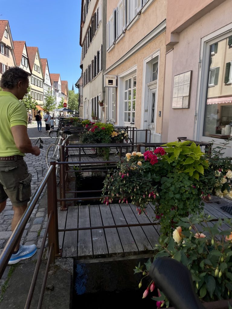 Blumengeschmückte Holzstege über einem Wasserlauf in der Altstadt von Tübingen, Deutschland. Historische Fachwerkhäuser mit roten Dächern säumen die Kopfsteinpflasterstraße, während Passanten durch die Fußgängerzone schlendern.