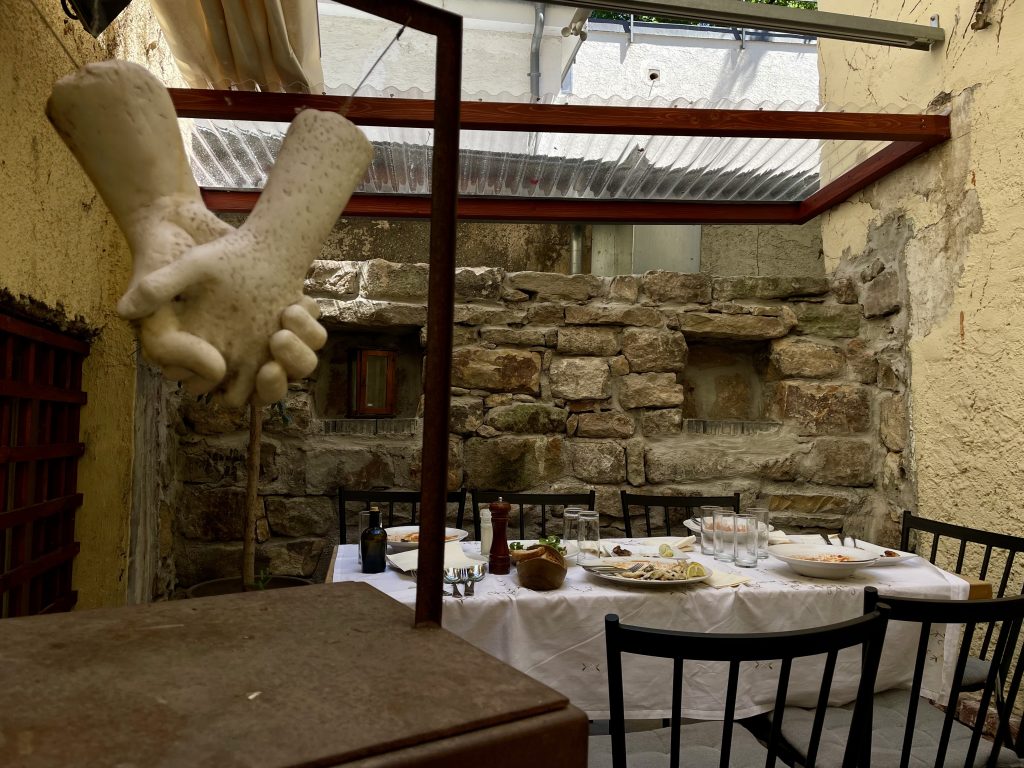 Versteckter Innenhof eines Restaurants in der Tübinger Altstadt mit skulpturalen Händehaltung, rustikaler Steinwand, gedecktem Tisch mit mediterranen Speisen – charmantes Zusammenspiel von Kunst, Kulinarik und Geschichte.