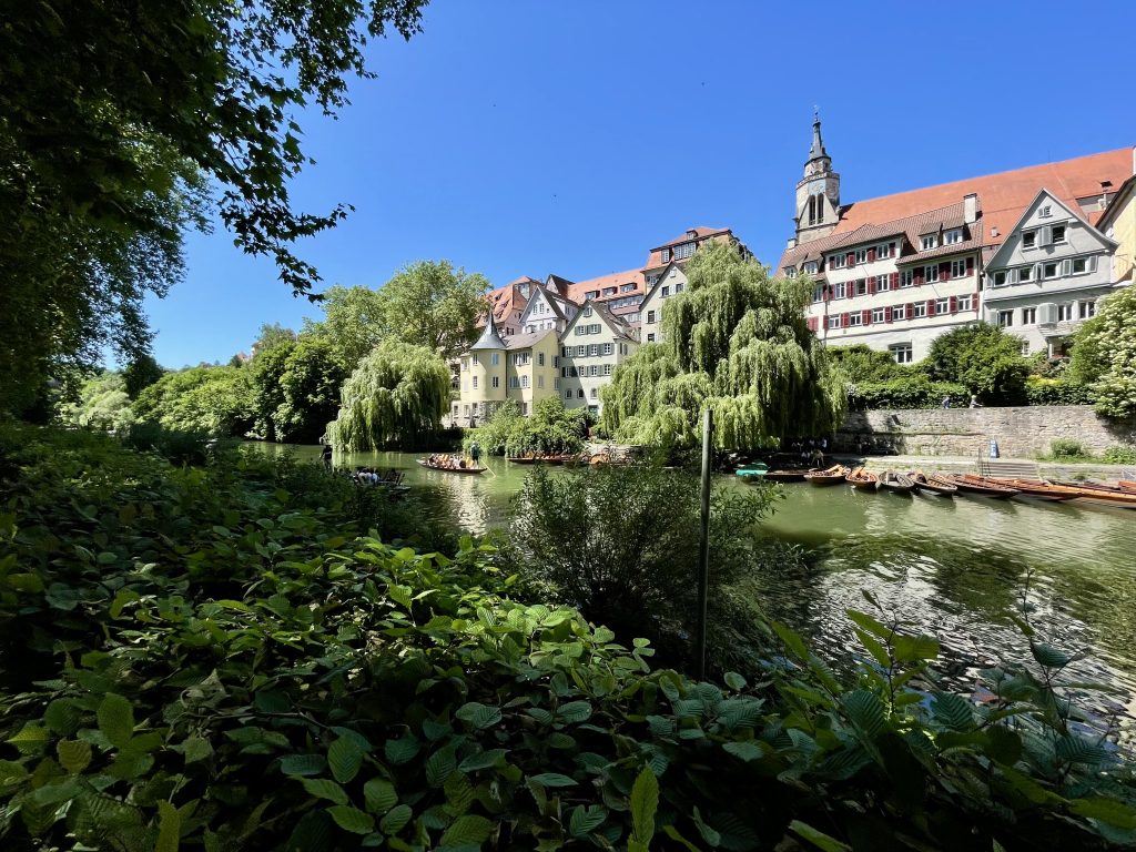Blick auf den Hölderlinturm am Neckar in Tübingen, gelbes historisches Haus mit Turm, umgeben von Weidenbäumen und Fachwerkhäusern; im Vordergrund Stocherkähne auf dem Fluss bei sonnigem Wetter.