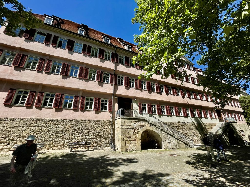 Historisches Gebäude der Alten Burse in Tübingen mit roter Fassade, Fensterläden und Freitreppe, einst Studentenwohnheim der Universität, umgeben von Bäumen und Kopfsteinpflaster.