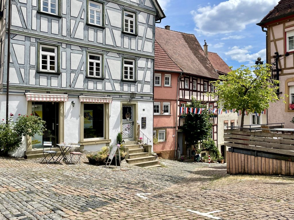 Historische Fachwerkhäuser an einem gepflasterten Altstadtplatz mit kleiner Außengastronomie, bunter Wimpelgirlande und sommerlicher Atmosphäre.