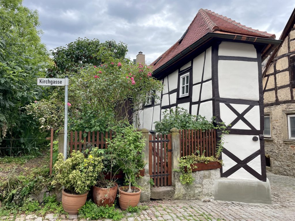 Fachwerkhaus in der Kirchgasse in Bad Wimpfen, umgeben von blühenden Rosensträuchern und Pflanzenkübeln, vor Kopfsteinpflaster und unter bewölktem Himmel.