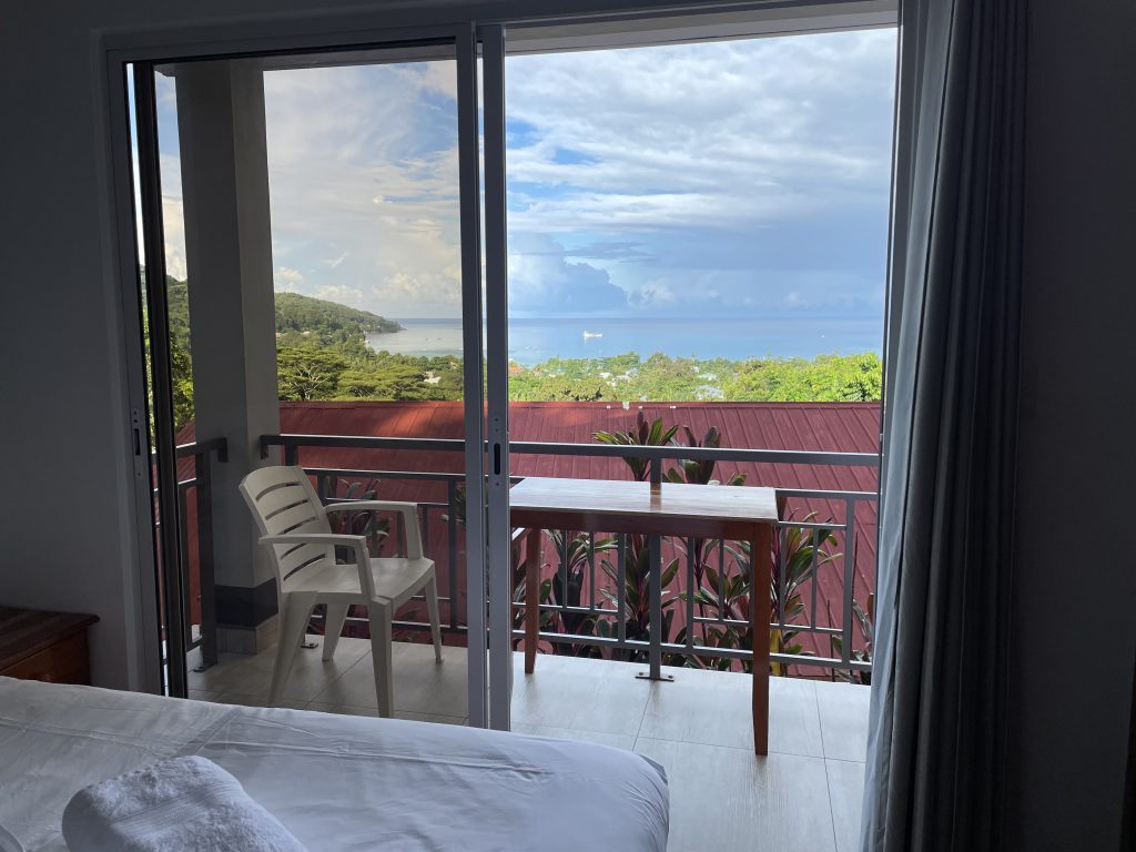 Ausblick aus einer Balkontüre. Auf dem Balkon steht ein Tisch und ein Stuhl. Als Ausblick sieht manBäume und das Meer. Der Himmel ist blau mit weißen Wolken durchzogen