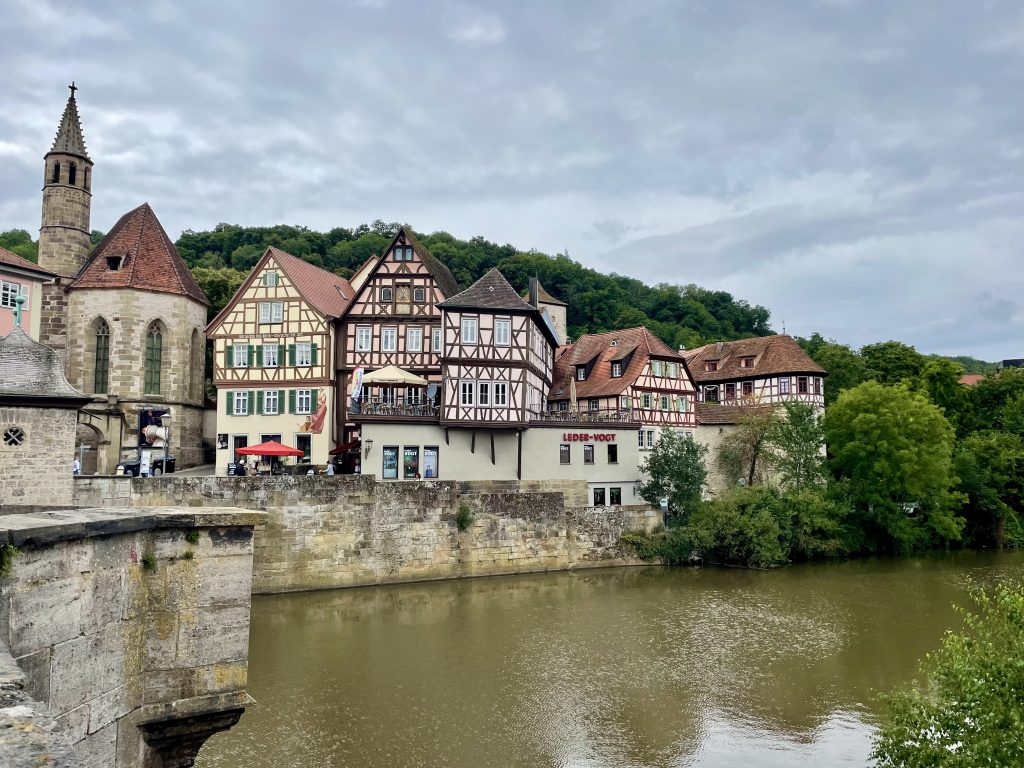 Ansicht auf Schwäbisch Hall im Juli. Vorne der Fluss und dann eine Kirche links sowie 5 Fachwerkhäuser. Wald it im Hintergrund sichtbar.