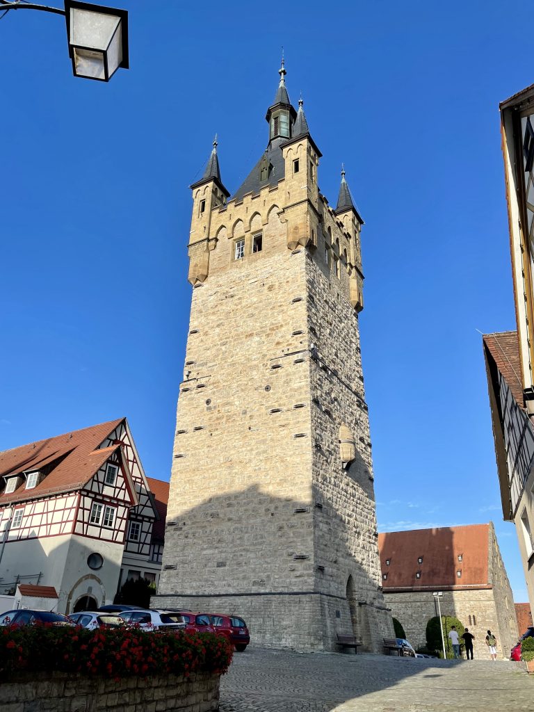 Der Blaue Turm in Bad Wimpfen, historischer Wachturm aus hellem Sandstein mit markanten Spitzdächern, vor blauem Himmel und umgeben von Fachwerkhäusern.“