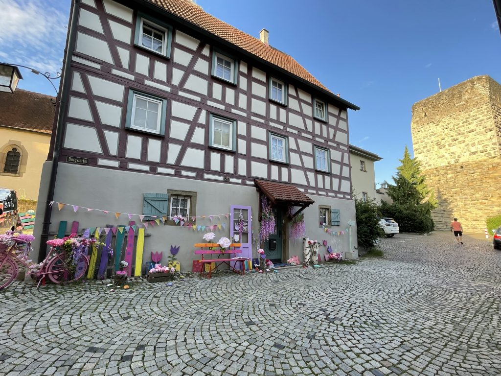 Blick in die Kirchgasse in Bad Wimpfen mit Fachwerkhäusern im Vordergrund, blühenden Rosensträuchern und dem Roten Turm aus Sandstein im Hintergrund