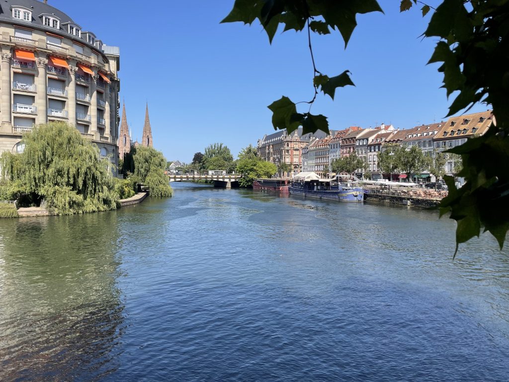 Blick auf die Ill in Straßburg mit historischen Fachwerkhäusern, Uferpromenade und Ausflugsschiffen – malerisches Reiseziel im Elsass.“