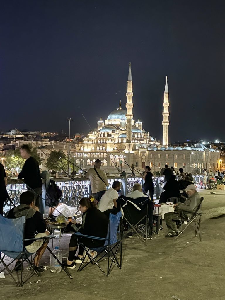 Beleuchtete Moschee in Istanbul bei Nacht, gesehen von der Galata-Brücke mit Menschen, die am Ufer sitzen, angeln und die Aussicht genießen.