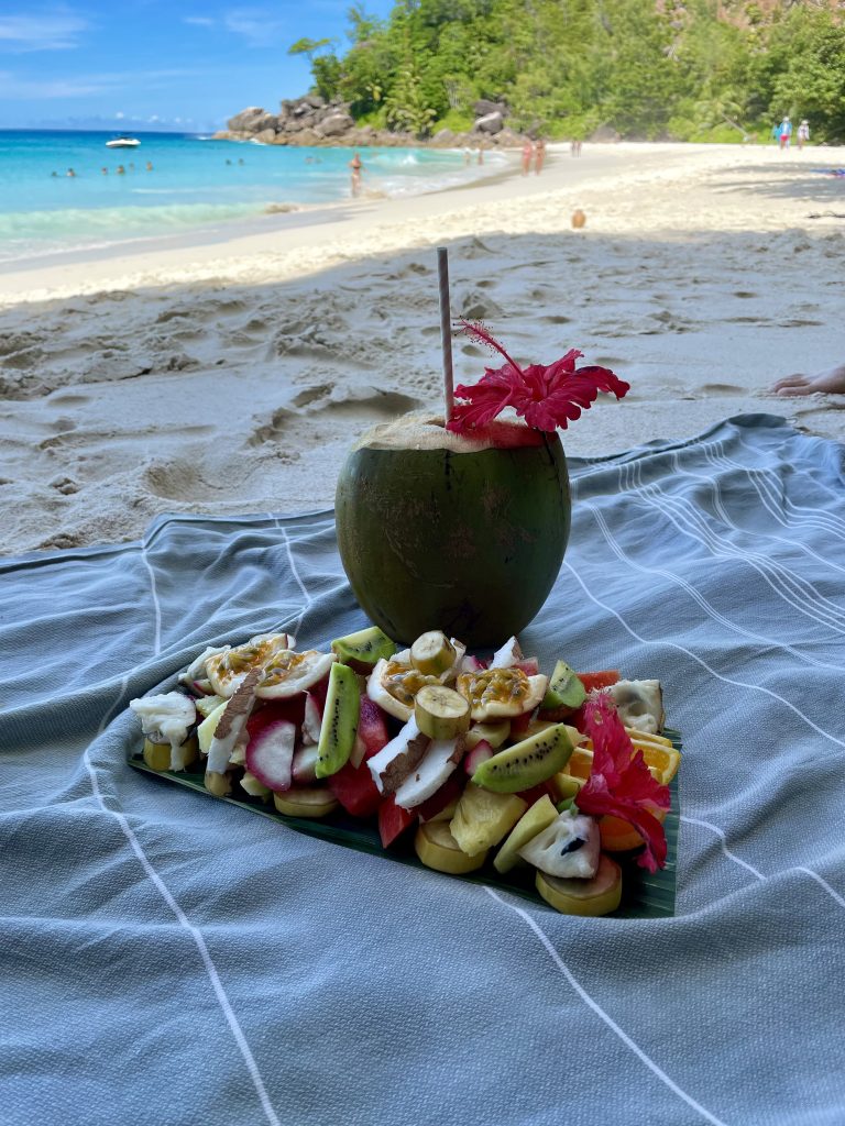 Auf einem Handtuch am Strand ist eingroßer Tellerst den verschiedensten Früchten und eine frische geöffnete Kokosnuss mit einer roten Blüte als Deko