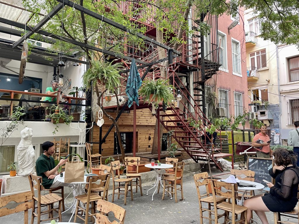Gemütliches Café mit Holzstühlen und Tischen in einem Innenhof in Istanbul, umgeben von Pflanzen, kunstvollen Dekorationen und einer roten Metalltreppe, mit Gästen, die Getränke genießen