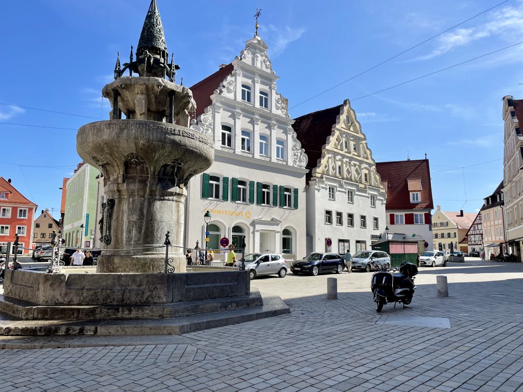 Der Marktplatz in Nördlingen. Im Vordergrund steht ein verzerrter Brunnen und im Hintergrund schöne Häuser, deren Giebel sehr kunstvoll verziert sind. Blauer Himmel mit wenigen weißen Schleierwolken. Auf dem Platz steht ein Vespa-Roller