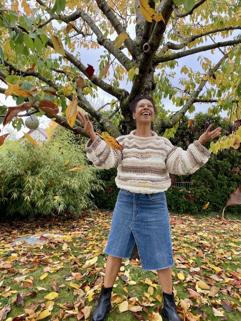 Frau steht untere einem Baum dessen Blätter sich schon in gelb und braun gefärbt hat. Sie hat einen selbst gestrickten Pullover in vielen beigetönen an und einen asymmetrisch geschnittenen Jeansrock an. Sie wirf Blätter in die Luft