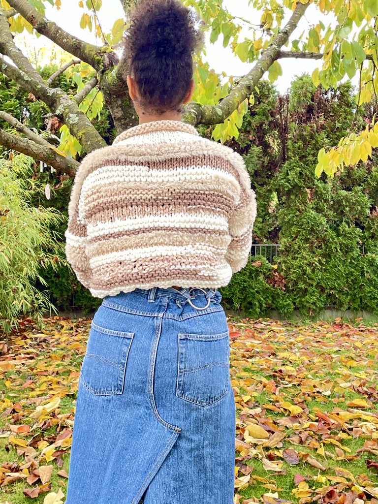 Frau von hinten mit einem Wollpullover und Jeansrock an steht unter einem Baum in Herbstfarben