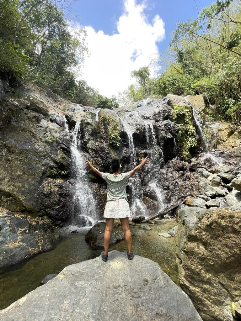Frau steht mit ausgebreiteten Armen vor einem Wasserfall auf Tobago