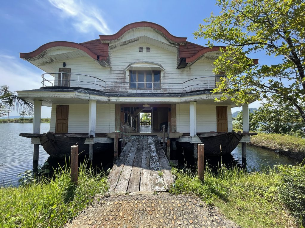 Ein Lost Place auf Koh Chang das Boat Chalet. im Vordergrund steht ein großes verwahrlostes Gebäude auf das ein Holzsteg führt. Rundherum ist Wasser und etwas Wiese.
