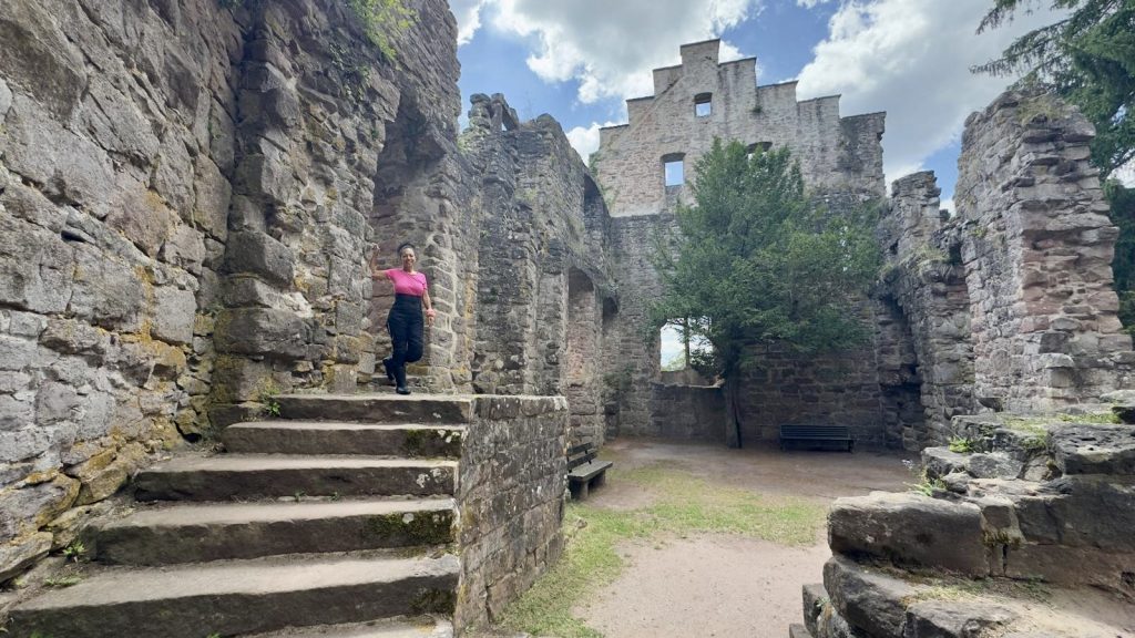 Frau in pinkem Shirt und schwarzer Hose steht auf einer Steintreppe in den Ruinen einer mittelalterlichen Burg bei sonnigem Wetter – historische Mauern, gotische Fensterbögen und ein Baum im Innenhof schaffen eine malerische Kulisse.